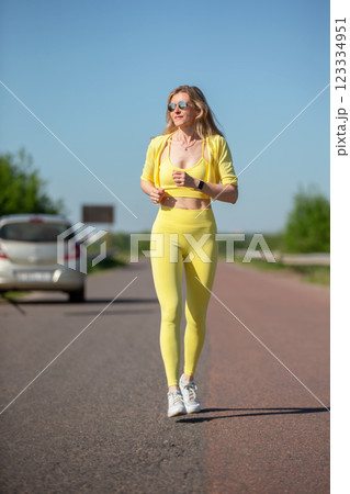 A woman in yellow running clothes is walking down a road. She is wearing a watch and sunglasses 123334951