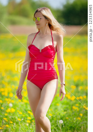 Beautiful adult blonde woman in a red swimsuit against the background of a meadow. 123334965