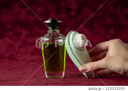 red burgundy velvet background there is a glass jar with green massage oil, on which a gently mint vacuum vibrator is leaning color device for girls relaxation for adults female hand takes a vibrator 123335205