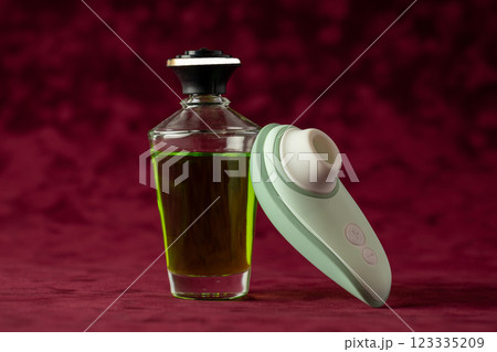 On a red burgundy velvet background there is a glass jar with green massage oil, on which a gently mint vacuum vibrator is leaning color device for girls, relaxation for adults On a red burgundy velvet background there is a glass jar with green massage oil, on which a gently mint vacuum vibrator is leaning color device for girls, relaxation for adults 123335209