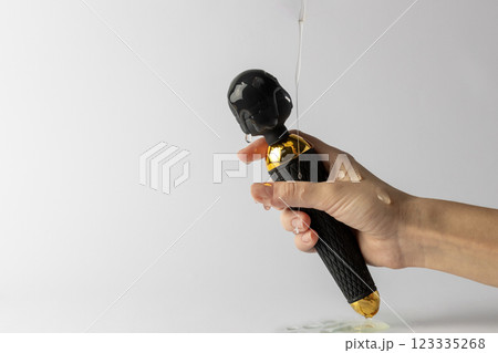 white background woman's hand with lubricant on her fingers holds a black large vibrator in the shape of a microphone doused with transparent water lubricant sexual pleasure toys for adults 123335268