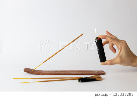 close-up on white background on a long brown stand with stars for incense sticks a new beige palosanto meditation stick is placed a hand with a black lighter setting fire 123335295
