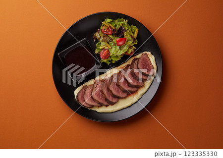 on orange background on a rectangular black plate lies mashed potatoes on which pieces of filet mignon are laid out, served with mushrooms and tomato on orange background on a rectangular black plate lies mashed potatoes on which pieces of filet mignon are laid out, served with mushrooms and tomato 123335330