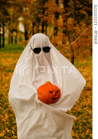 Woman in white sheet holding jack o'lantern against autumn forest background. Halloween costume. Vertical photo. Woman in white sheet holding jack o'lantern against autumn forest background. Halloween costume. Vertical photo. 123335570