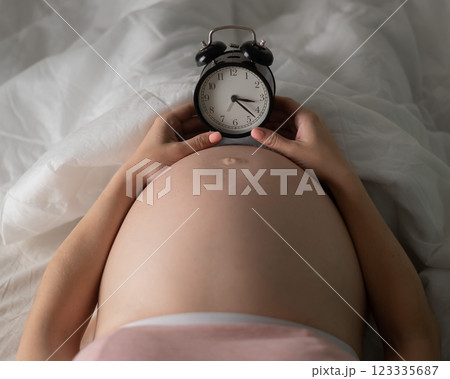 A pregnant woman lies in bed and holds an alarm clock. Close up of the belly. 123335687
