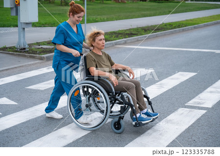 Red-haired nurse pushing an elderly woman in a wheelchair across the road. Red-haired nurse pushing an elderly woman in a wheelchair across the road. 123335788