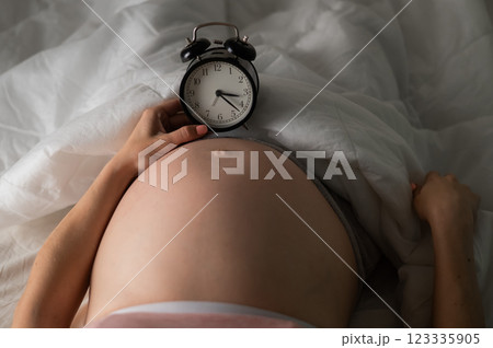 A pregnant woman lies in bed and holds an alarm clock. Close up of the belly. A pregnant woman lies in bed and holds an alarm clock. Close up of the belly. 123335905