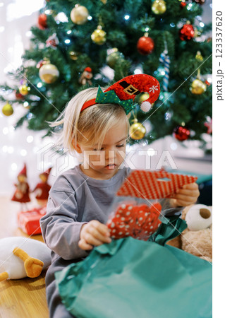 Little girl in a hoop with a gnome hat examines a package with red bows sitting by the Christmas tree 123337620
