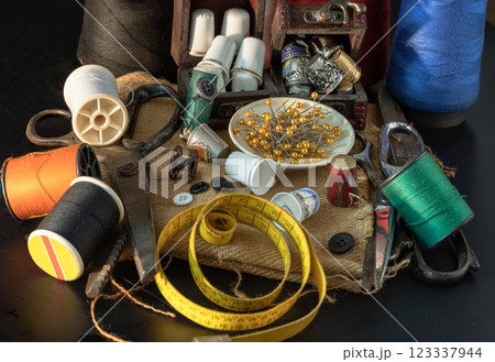 Spools of thread of various colors, Thimbles in small wooden treasure chest, Steel scissors, Sewing needles stuck in a green miniature spools thread and Measuring tape including pins in small round 123337944