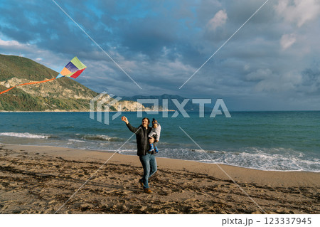 Dad with a little girl in his arms looks at a hovering kite, walking with it along the seashore 123337945