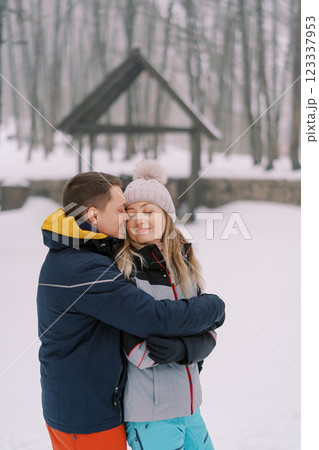Smiling man hugging woman from behind, touching her temple with his nose in snowy park 123337953