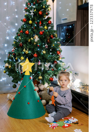 Little girl holds a felt toy in her hands while sitting on the floor near a toy Christmas tree Little girl holds a felt toy in her hands while sitting on the floor near a toy Christmas tree 123337978
