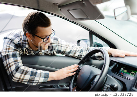Young man looking through window inside new car, checking it before purchase at modern dealership store. Cheerful guy buying or renting automobile at showroom 123338729