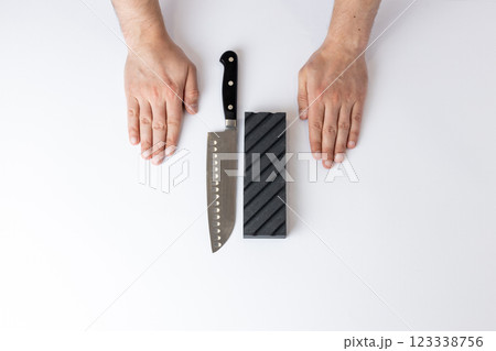 on white background on the table there lies a large knife for sharpening and a black grooved sharpening stone on the first floor, men's hands lie next to it parallel to the knife on white background on the table there lies a large knife for sharpening and a black grooved sharpening stone on the first floor, men's hands lie next to it parallel to the knife 123338756