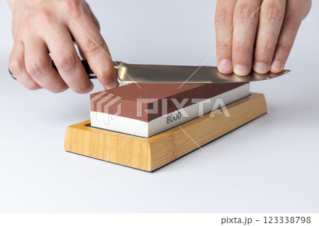 close up on white background on a table, man hands sharpen a steel knife protection against cuts, a red medium-hard stone for sharpening knives 123338798