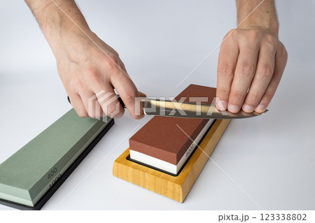 close up on white background on a table, man hands sharpen a steel knife protection against cuts, a red medium-hard stone for sharpening knives a green whetstone lies nearby 123338802