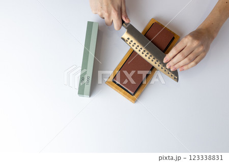 top shot on white background on a table, women's hands sharpen a steel knife, a lock under the arm, protection against cuts, a red medium-hard stone for sharpening knives green whetstone nearby top shot on white background on a table, women's hands sharpen a steel knife, a lock under the arm, protection against cuts, a red medium-hard stone for sharpening knives green whetstone nearby 123338831