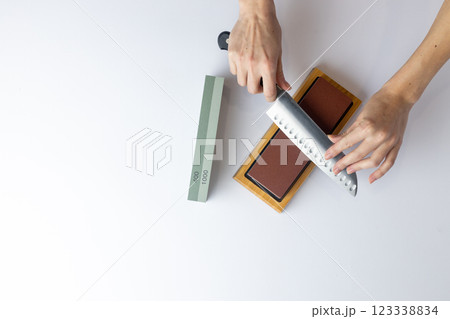 top shot on white background on a table, women's hands sharpen a steel knife, a lock under the arm, protection against cuts, a red medium-hard stone for sharpening knives green whetstone nearby top shot on white background on a table, women's hands sharpen a steel knife, a lock under the arm, protection against cuts, a red medium-hard stone for sharpening knives green whetstone nearby 123338834
