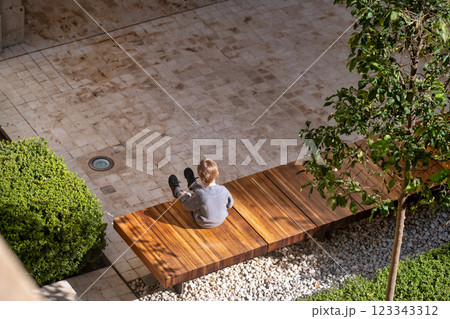 The image features an overhead view of an outdoor setting with a person sitting on a wooden bench. The person appears to be wearing light-colored clothes. The bench is surrounded by a neatly The image features an overhead view of an outdoor setting with a person sitting on a wooden bench. The person appears to be wearing light-colored clothes. The bench is surrounded by a neatly 123343312