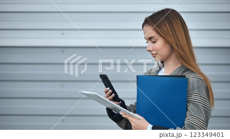 Young businesswoman holding folder and using digital tablet and smartphone, standing outdoors near modern office building Young businesswoman holding folder and using digital tablet and smartphone, standing outdoors near modern office building 123349401