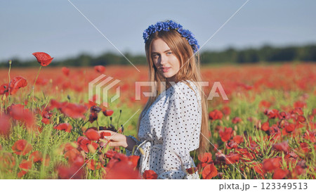 Blonde woman in white polka dot dress, blue floral headpiece, caressing red poppies under bright summer sunlight 123349513