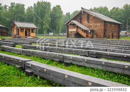 Rows of weathered wooden benches occupy the foreground, facing two traditional russian log houses under a cloudy sky 123349832