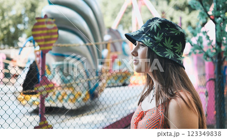 Girl wearing a bucket hat with cannabis leaf prints looking at an amusement park ride in summer 123349838