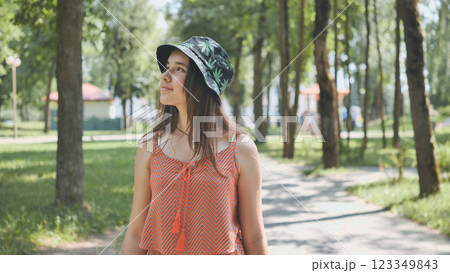 Young female sporting trendy bucket hat, bright orange top, strolling cheerfully across sunlit park path, embracing summer's carefree energy Young female sporting trendy bucket hat, bright orange top, strolling cheerfully across sunlit park path, embracing summer's carefree energy 123349843