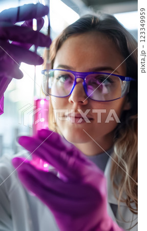 focused female scientist in glasses and purple gloves holds up test tube with biotech sample liquid, biotechnology experiment in laboratory environment 123349959