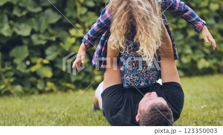 Father lying on grass, lifting young daughter overhead, bonding during playful backyard moment on bright summer day 123350003