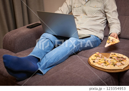 man sitting on sofa and eating pizza while working on laptop, close-up man sitting on sofa and eating pizza while working on laptop, close-up 123354953