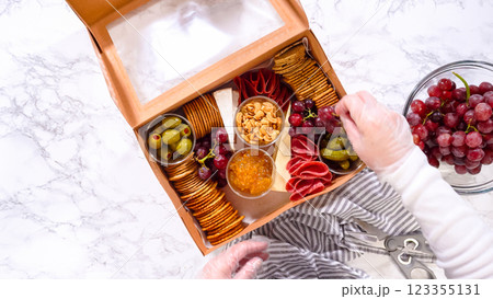 Flat lay. Hands are meticulously adding fresh red grapes to a bowl, complementing a beautifully arranged charcuterie box brimming with a variety of cheeses, olives, and cured meats, set against a Flat lay. Hands are meticulously adding fresh red grapes to a bowl, complementing a beautifully arranged charcuterie box brimming with a variety of cheeses, olives, and cured meats, set against a 123355131
