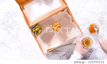 Flat lay. Hands are meticulously adding fresh red grapes to a bowl, complementing a beautifully arranged charcuterie box brimming with a variety of cheeses, olives, and cured meats, set against a 123355152