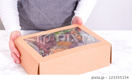 Hands are meticulously adding fresh red grapes to a bowl, complementing a beautifully arranged charcuterie box brimming with a variety of cheeses, olives, and cured meats, set against a sleek marble Hands are meticulously adding fresh red grapes to a bowl, complementing a beautifully arranged charcuterie box brimming with a variety of cheeses, olives, and cured meats, set against a sleek marble 123355154