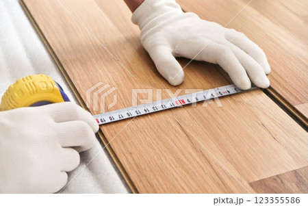 Installing laminated floor, detail on man hands in white gloves holding measuring tape over wooden tile Installing laminated floor, detail on man hands in white gloves holding measuring tape over wooden tile 123355586