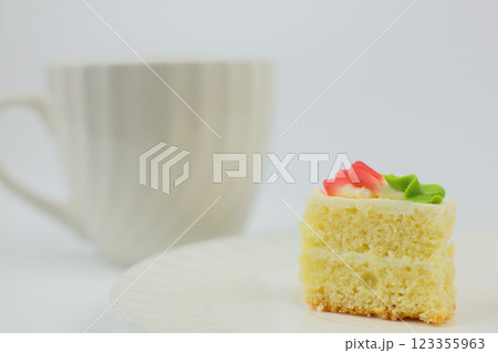 A cake consisting of an airy sponge cake and white cream, decorated with a mastic flower on top, stands on a white saucer in close-up against the background of a white mug with a hot drink. The A cake consisting of an airy sponge cake and white cream, decorated with a mastic flower on top, stands on a white saucer in close-up against the background of a white mug with a hot drink. The 123355963