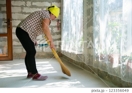 A woman in a yellow headscarf sweeps the floor of a building using a straw broom. The floor is light grey and a window is in the background. A woman in a yellow headscarf sweeps the floor of a building using a straw broom. The floor is light grey and a window is in the background. 123356049
