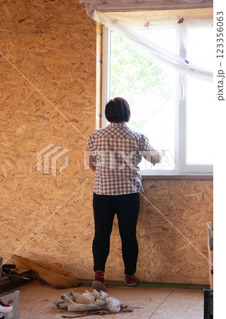 Woman installs a bright, white window in a wood paneled room with curtains. She is wearing black pants, a plaid shirt and red shoes. 123356063
