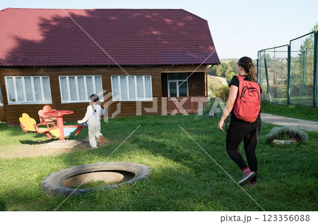 Child playing on merry-go-round while woman walks in a grassy yard near a rustic wooden house on a sunny day at a park or school. 123356088