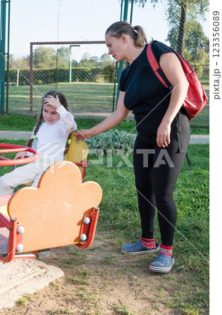 A young girl on a merry-go-round looks distressed as a woman with a backpack stands nearby on the grass in an outdoor park setting. A young girl on a merry-go-round looks distressed as a woman with a backpack stands nearby on the grass in an outdoor park setting. 123356089