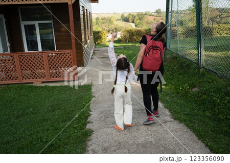 A child points upward while holding hands with an adult, walking on a path by a house and green landscape, enjoying a sunny day. A child points upward while holding hands with an adult, walking on a path by a house and green landscape, enjoying a sunny day. 123356090