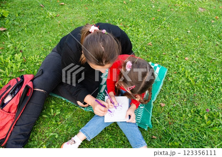 A woman and a child are drawing together outdoors on a blanket. They are enjoying a sunny day on the green grass. A woman and a child are drawing together outdoors on a blanket. They are enjoying a sunny day on the green grass. 123356111