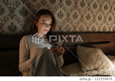Attractive redhead young woman relaxing using smartphone sitting in bed at night, looking to screen, enjoying social media and online communication in cozy darkness of bedroom. Attractive redhead young woman relaxing using smartphone sitting in bed at night, looking to screen, enjoying social media and online communication in cozy darkness of bedroom. 123359878