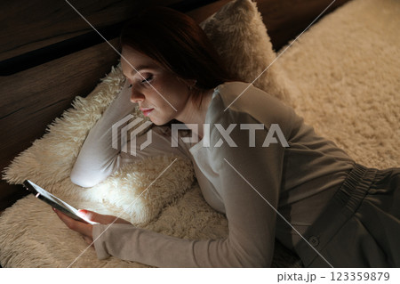 Top view of tired young woman lying in bed at night, using smartphone to connect with friends and enjoy social media, screen warm glow illuminating face, creating cozy atmosphere in dark bedroom Top view of tired young woman lying in bed at night, using smartphone to connect with friends and enjoy social media, screen warm glow illuminating face, creating cozy atmosphere in dark bedroom 123359879