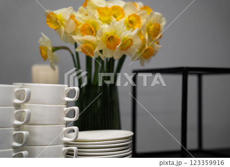 Many clean white cups and plates on the table near the coffee maker and vase with daffodils 123359916