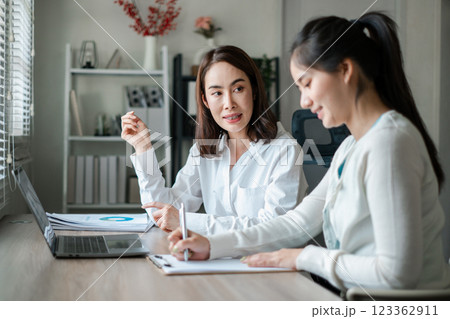 Two women working together in a bright office, discussing documents and using a laptop, showcasing teamwork and collaboration. Two women working together in a bright office, discussing documents and using a laptop, showcasing teamwork and collaboration. 123362911