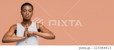 African American young woman wearing a striped tank top practices yoga with her hands in front of her chest, in a meditative pose. She is in front of a peach-colored background. 123364411