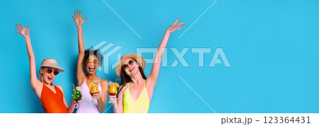 Three women wearing swimwear and sunglasses stand together in front of a blue background. They have their arms raised in the air, and each woman is holding a cocktail drink, copy space Three women wearing swimwear and sunglasses stand together in front of a blue background. They have their arms raised in the air, and each woman is holding a cocktail drink, copy space 123364431