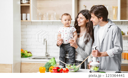 A happy family is preparing a healthy meal together in their modern kitchen. The mother is holding their baby, who is looking at the camera with a curious expression, copy space 123364441