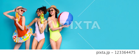 This image shows three women in swimsuits posing against a blue background. They are holding beach toys, including a beach ball and water guns. The women are smiling and appear to be having fun. This image shows three women in swimsuits posing against a blue background. They are holding beach toys, including a beach ball and water guns. The women are smiling and appear to be having fun. 123364475